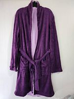 Full view of purple toweling bathrobe on hanger showing overall shape, belt, front pockets, and long sleeves.