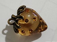 Front angled close-up of the strawberry pendant showing clear smoky glass with golden-brown internal dots and brass-tone leaf loop attachment.