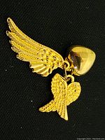 Clear close-up photo of gold pendant with a large wing charm, smaller wing charm, and heart charm on a black background.