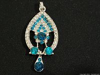 Front view of silver-tone pendant with turquoise blue and clear rhinestone faux diamonds on black background.