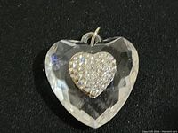 Close-up photo of clear heart-shaped pendant with rhinestone encrusted smaller heart in center, showing faceted edges and metal loop for attachment.