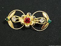 Close-up photo of gold tone decorative pin with red center gem, clear stones, and small green stones on leaf shapes, on black background.