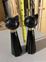 Two black glass cat-shaped cologne bottles standing upright, measured with tape showing about 8 inches tall.