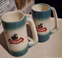 Photo of two ceramic O'Keefe Beer mugs showing their shape, handle, and logo featuring a horse and shield with gradient blue and white coloring.