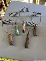 Five vintage potato mashers lying on a flat surface, showing wooden handles and metal mashing heads of different designs and wear.
