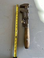 Full view of antique adjustable wrench next to measuring tape showing approximate length of 15 inches. Wooden handle and metal jaw visible.