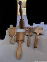 Top view of four wooden butter paddles showing different flat pad shapes and grooved versus smooth faces, alongside one wooden pestle standing upright behind them