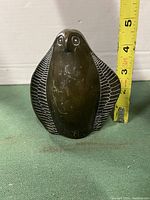 Front view of carved stone penguin beside tape measure showing height around 3.5 inches. Detailed textured wings and round eyes visible.