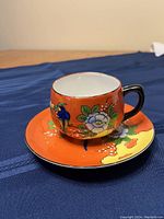 Cup and saucer shown together on blue fabric background highlighting detailed hand-painted bird and flower artwork on an orange base.