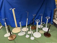 Lot of 14 doll display stands, variety of sizes and base materials, mostly metal with some wooden bases, all with adjustable wire holders