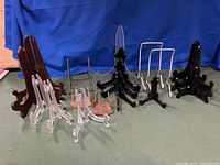 Photo showing 18 collector plate display stands in various materials and sizes, including clear acrylic, dark wood, black plastic, metal wire, and small brown wood stands arranged on a flat surface against a blue backdrop.
