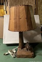 Front view of the lamp showing the wooden slat lampshade, carved tree trunk style lamp stand, and the bear cub figurine attached to the base, with power cord coiled beside.