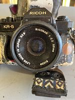 Close-up front view showing Ricoh KR-5 camera with prominent Ricoh Riconar 55mm lens marked 'Lens Made in Japan'. Strap visible below.