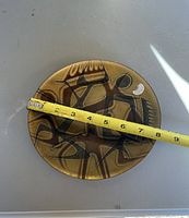 Top side of the round glazed tin dish showing an indigenous style graphic pattern in browns and blacks, measured with a tape around 8 inches diameter.