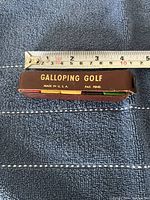Photo of the brown leather case with gold embossed text 'Galloping Golf Made in U.S.A. Pat. Pend.' placed on a dark blue fabric background with a measuring tape showing approximately 5 inches in length.