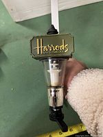 Close-up of the Harrod's Knightsbridge branded top section of the liquor bottle dispenser showing the transparent measuring chamber and dispensing lever.