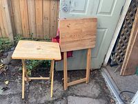 Two foldable wooden TV dinner tables, one standing folded against a door, the other opened showing the tabletop and legs.
