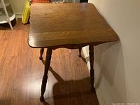 Square antique wooden table on a hardwood floor against a wall, visible tabletop and legs showing wear and wood grain.