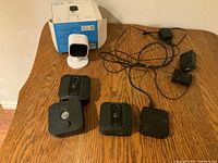 Three black Blink XT2 outdoor cameras with motion sensors and one power adapter on wooden table alongside a white Blink Mini camera in front of its box.