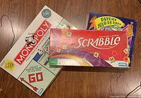 Photo shows Monopoly, Payday, and Scrabble board games in original boxes stacked on a wooden floor.