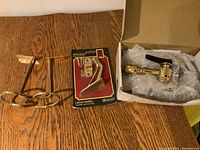 Three brass hardware items: a faucet-style sink hose sprayer with lever, a brass hook in original packaging, and two brass curtain holdbacks with double ring design, all shown on a wood surface and partially in a box with bubble wrap.