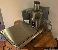 Juicer and grill appliances on a wooden table showing stainless steel juicer with pulp container and clear juice jug, alongside the stainless steel grill closed and resting sideways.