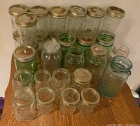 Over two dozen varied canning jars arranged on a wooden surface, including different sizes and colors with metal lids, showing signs of use.