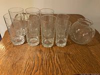 Full view of six large glasses, two smaller glasses grouped in front of a wooden table, alongside a spherical decorative glass vase with etched floral pattern.