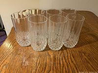 Seven clear crystal water glasses arranged in two rows on a wooden table with a visible textured pattern on the base of each glass.