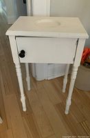 Front view of small antique bedside table painted off white. Shows turned legs, closed door with black knob, and circular indentation on top.