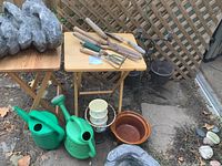 Garden tools, watering cans, and ceramic pots on a wooden table and on the ground.