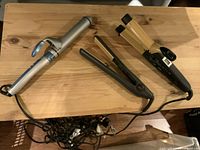 Three hair styling tools on wooden surface: BabyBliss Pro curling iron (silver/blue), Chi hair curler/straightener (black/gold), Chi hair straightener (black/beige)