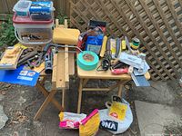 Wide view of various handyman tools and materials arranged on small folding tables outdoors. Includes containers of screws, caulk tubes, putty knives, tapes, gloves, adjustable wrench, spray can, utility knife, sponge, and cement bags underneath.