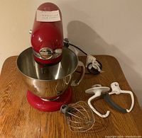 Front view of red mixer on wooden table with stainless steel bowl and three attachments laid out