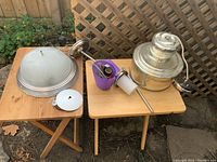 Five light fixtures displayed on two small wooden tables outdoors; includes a dome ceiling fixture, a metal pendant light, and a container with electrical parts.