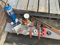 Lot layout on outdoor wooden steps showing plumbing tools and fittings including wrenches, pipe cutters, copper pipes, and torch.