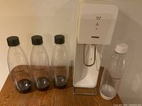 White SodaStream machine standing upright next to four clear plastic SodaStream bottles, one bottle has a white cap and appears brand new, others have black caps.