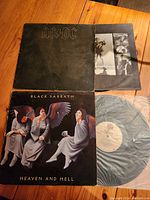 Two vinyl album covers and one vinyl record. Top album is AC/DC with a plain black cover. Bottom album is Black Sabbath 'Heaven and Hell' with illustrated cover. Black Sabbath record partially visible inside sleeve.