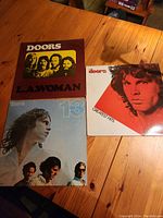 Photo showing three vinyl record albums by The Doors: L.A. Woman, 13, and Greatest Hits on a wooden surface.