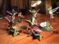 Five assorted hummingbird figurines arranged on a wooden surface, various sizes and perched on flowers or branches