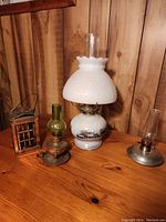 Four assorted lanterns on wood surface and wood paneled background: large white milk glass Currier and Ives style lamp, smaller green oil lamp, small clear glass oil lamp with metal base, decorative telephone booth metal lantern.
