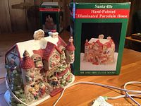 Hand-painted illuminated porcelain Christmas village house in front of its box, showing detailed Santa Claus themed decorations with figures and snow accents.