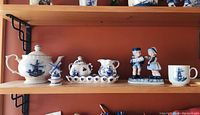 Shelf display of Delft blue porcelain items including teapot, cream and sugar set on tray, pair of couple figurines, and cup.