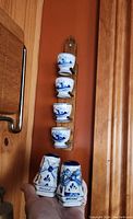 Four blue and white Delft ceramic egg cups mounted vertically on a wooden holder against an orange wall, accompanied by a pair of Delft salt and pepper shakers decorated with windmills, held in hand for scale.