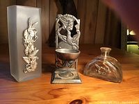 Photo of three assorted decorative pieces: frosted glass vase with pewter hummingbird applique, pewter hummingbird candle holder, and clear glass bottle with embossed hummingbird.