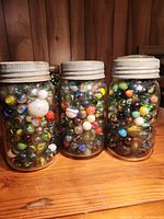 Three clear glass Canadian made mason jars filled with various colorful marbles on a wooden surface with visible metal lids showing wear.