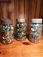 Three vintage glass jars filled with colorful marbles, placed on a wooden surface with a wood-panel background. Each jar has a different lid color and condition.