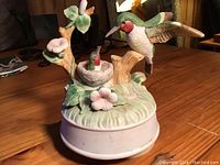 Porcelain or ceramic hummingbird music box on wooden surface; two hummingbirds with floral and nest details, round white base with slight wear marks.