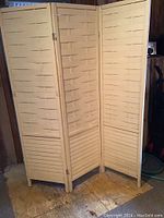 Photo shows a light-colored wooden room divider with three hinged panels featuring a woven slat design, folded partially and standing upright.
