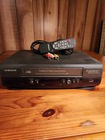 Front view of the Samsung VCR showing control buttons, model and system label, along with remote and AV cables on top.